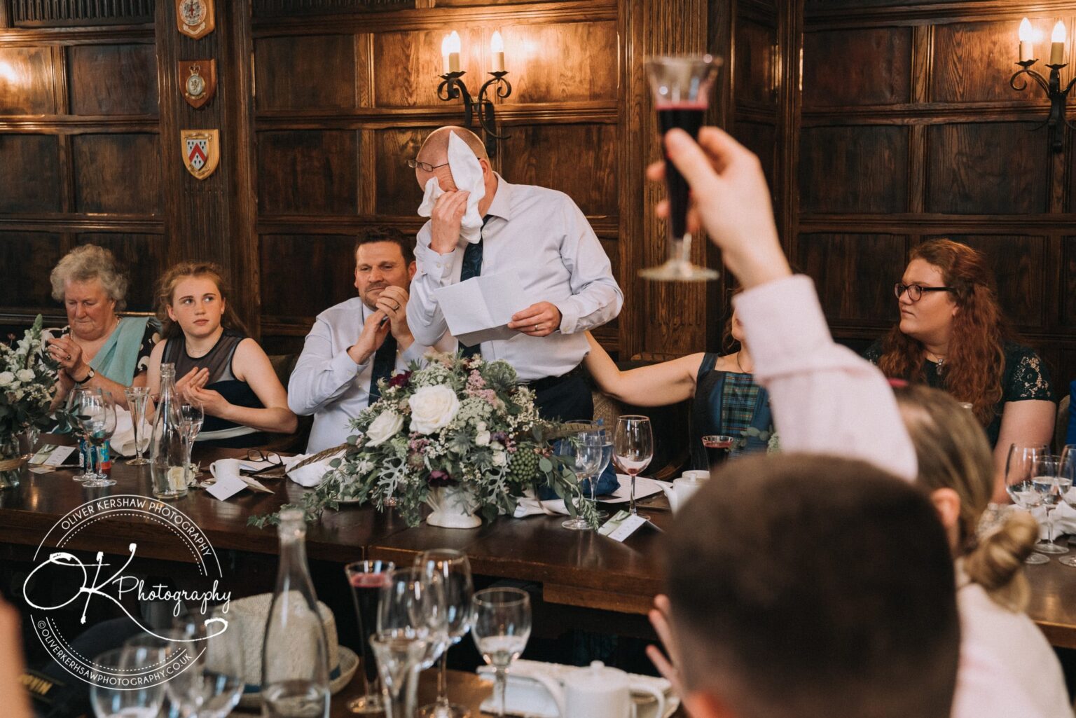 Wedding Photography George Hotel Stamford A group of people sitting at a table during a wedding reception, with one man standing and wiping his face with a napkin, while holding a speech.