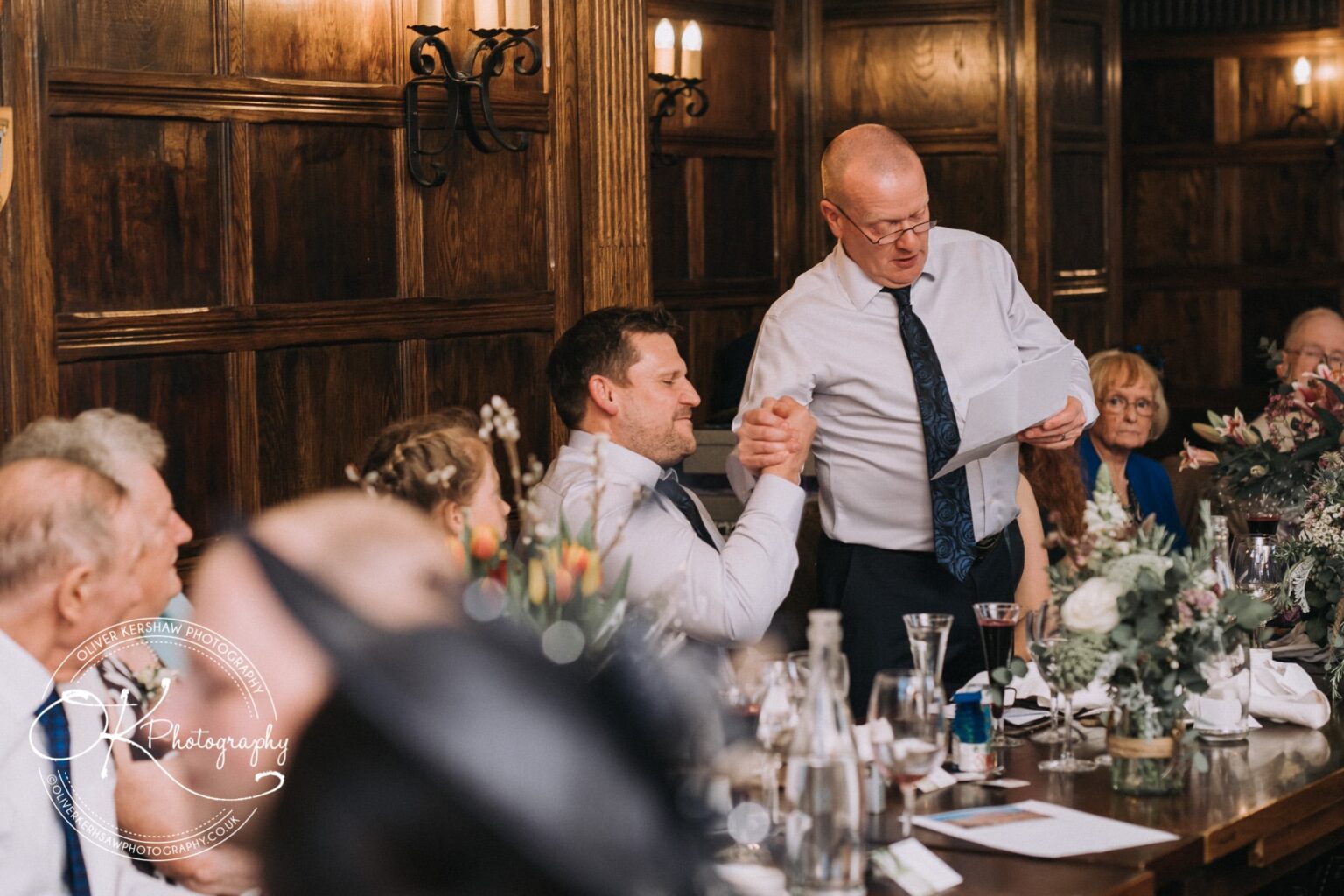 Wedding Photography George Hotel Stamford A man making a speech, shaking hands with another man at a dinner table in a wood-panelled room, with guests seated around.