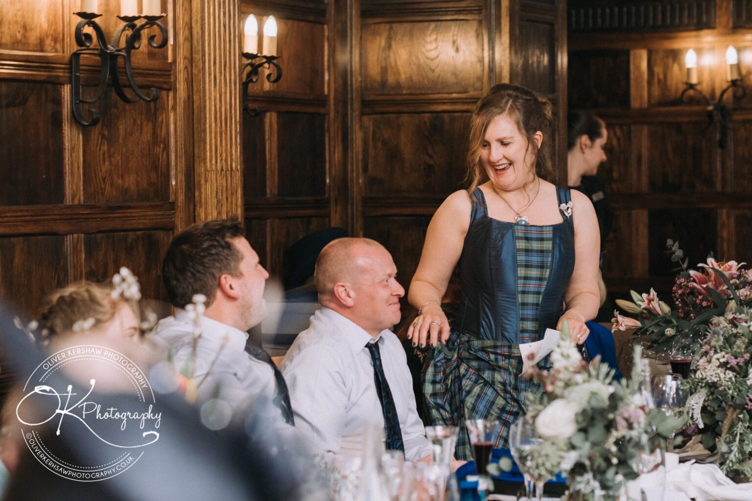 Wedding Photography George Hotel Stamford People sitting at a table, with a woman standing and smiling in a wood-paneled room decorated with flowers.