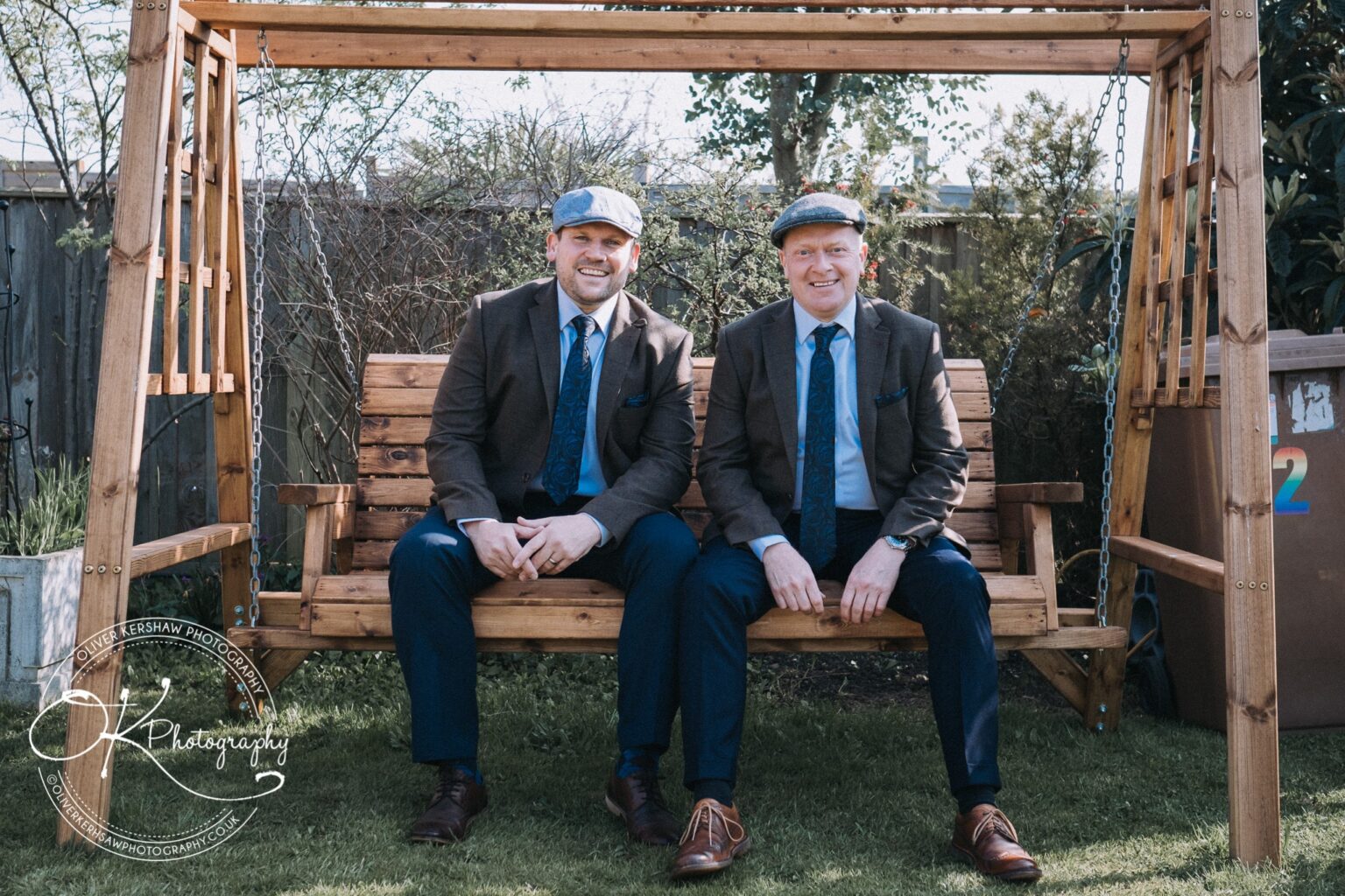 Wedding Photography George Hotel Stamford Two men sitting on a wooden garden swing, both wearing brown jackets, blue trousers, ties, and flat caps.