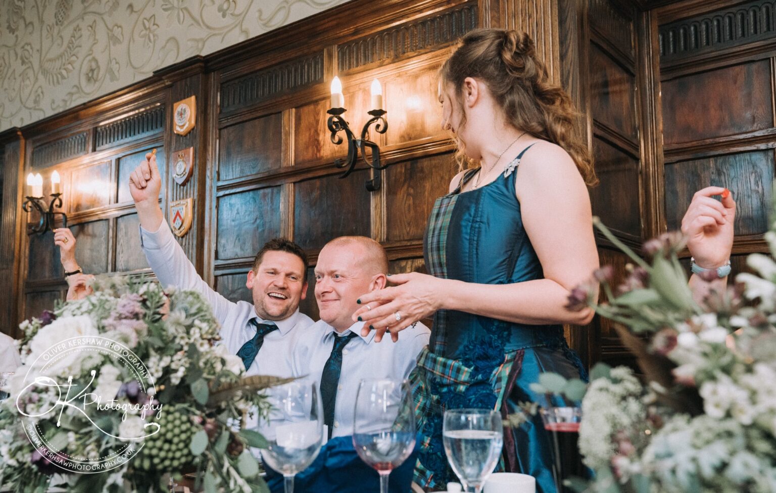 Wedding Photography George Hotel Stamford Guests in formal attire celebrating at a table with floral decorations, wooden wall panels, and candelabras in the background.