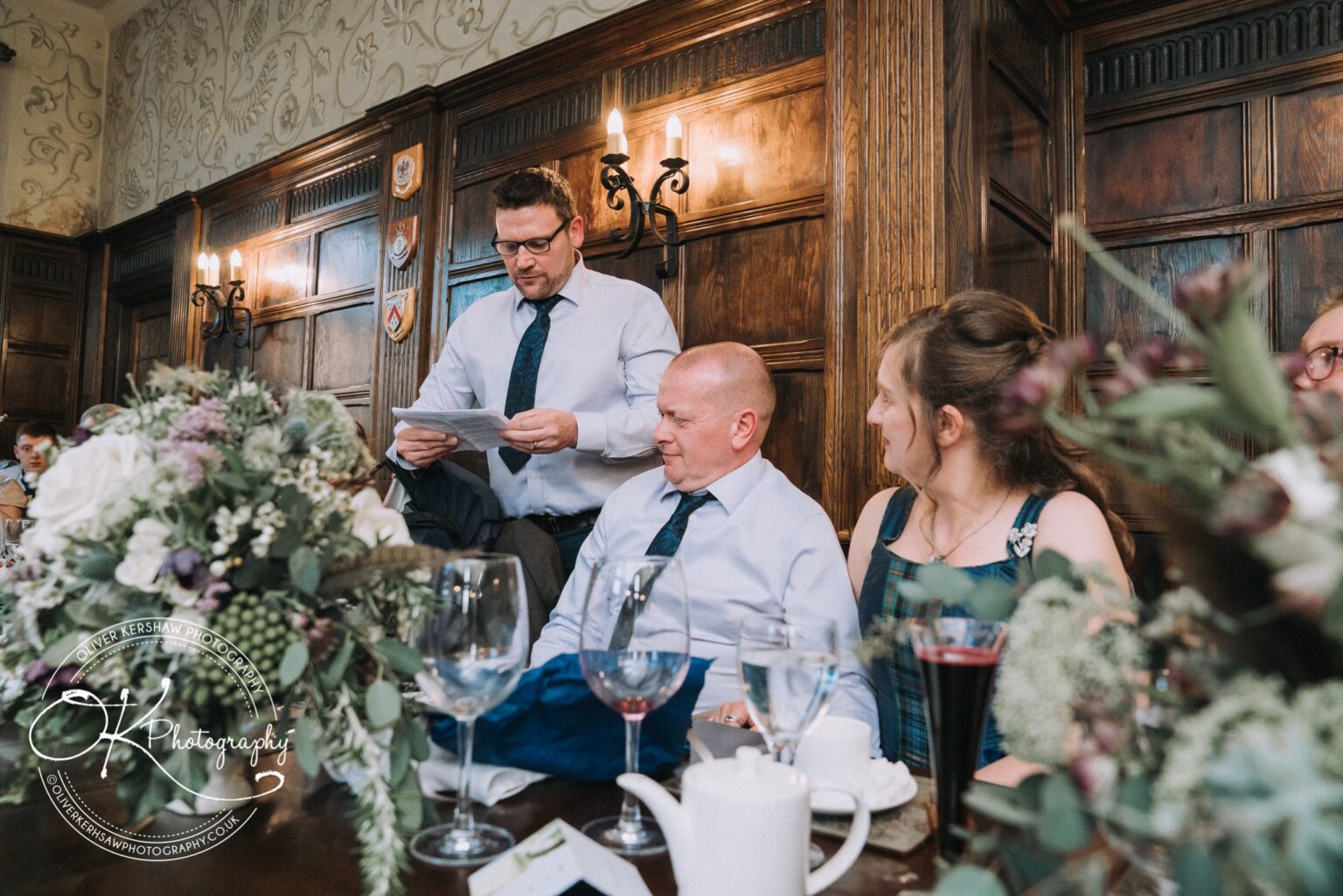Wedding Photography George Hotel Stamford A man standing and reading from a paper, with seated guests listening attentively at a table adorned with flowers and glasses, inside a wood-panelled room at George Hotel in Stamford.