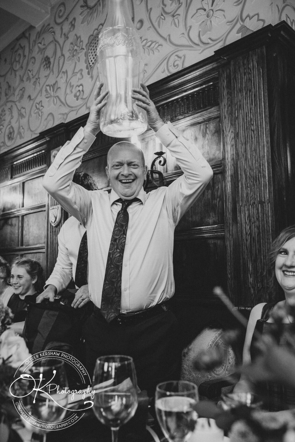 Wedding Photography George Hotel Stamford A man holding a large glass Coca-Cola bottle above his head at an event with other people smiling and seated around him.