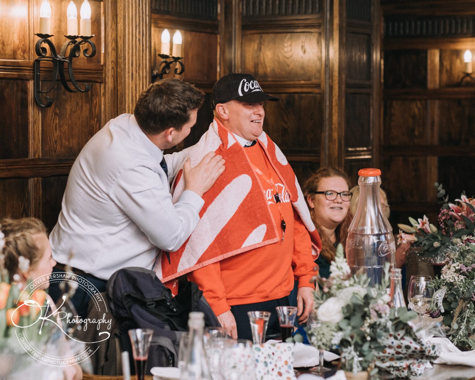 Wedding Photography George Hotel Stamford A man wearing a Coca-Cola hat and sweatshirt is being patted on the shoulder by another man in a white shirt, surrounded by people sitting at a dinner table in a wood-panelled room.