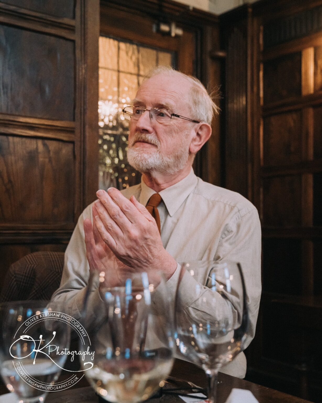 Wedding Photography George Hotel Stamford An elderly man in glasses and a light-coloured shirt clapping his hands while sitting at a table with wine glasses, in a wood-panelled room.