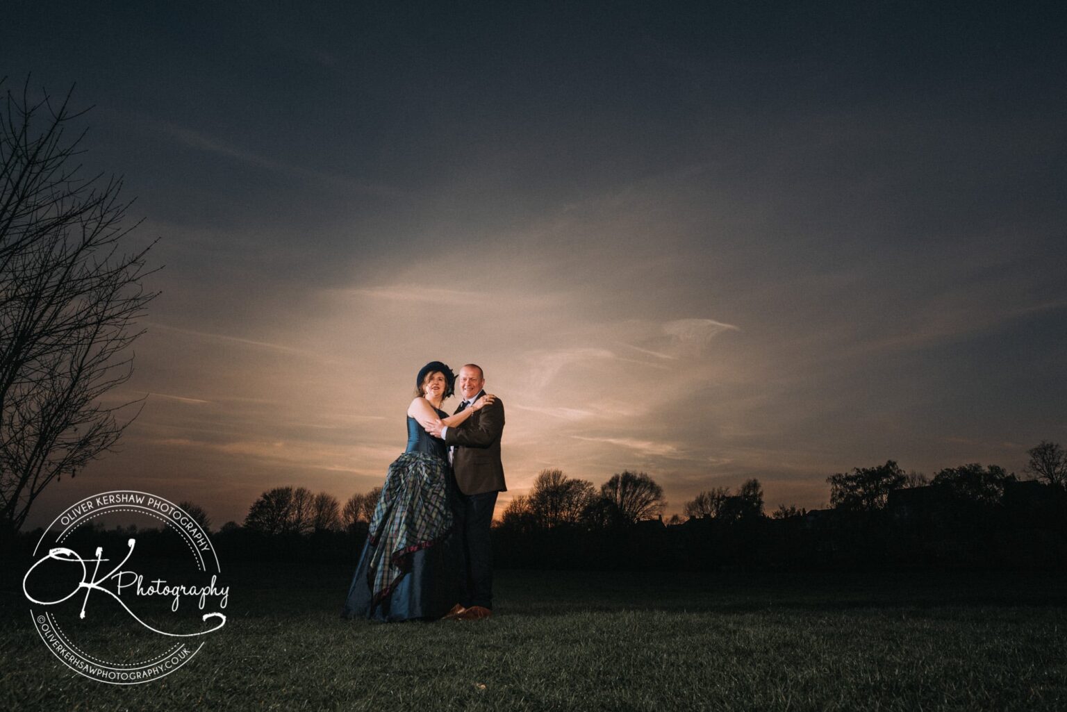 Wedding Photography George Hotel Stamford A couple in formal attire standing and embracing in an open field at sunset with trees in the background.