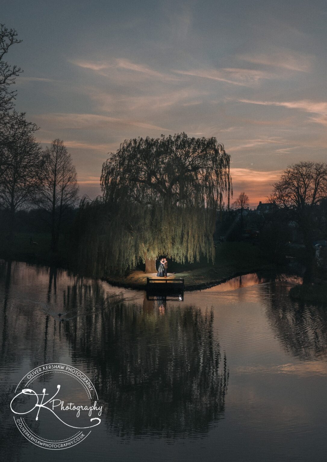 Wedding Photography George Hotel Stamford A couple stands on a pier by a weeping willow tree illuminated in the twilight at George Hotel in Stamford, with a reflective pond and a sunset sky in the background.