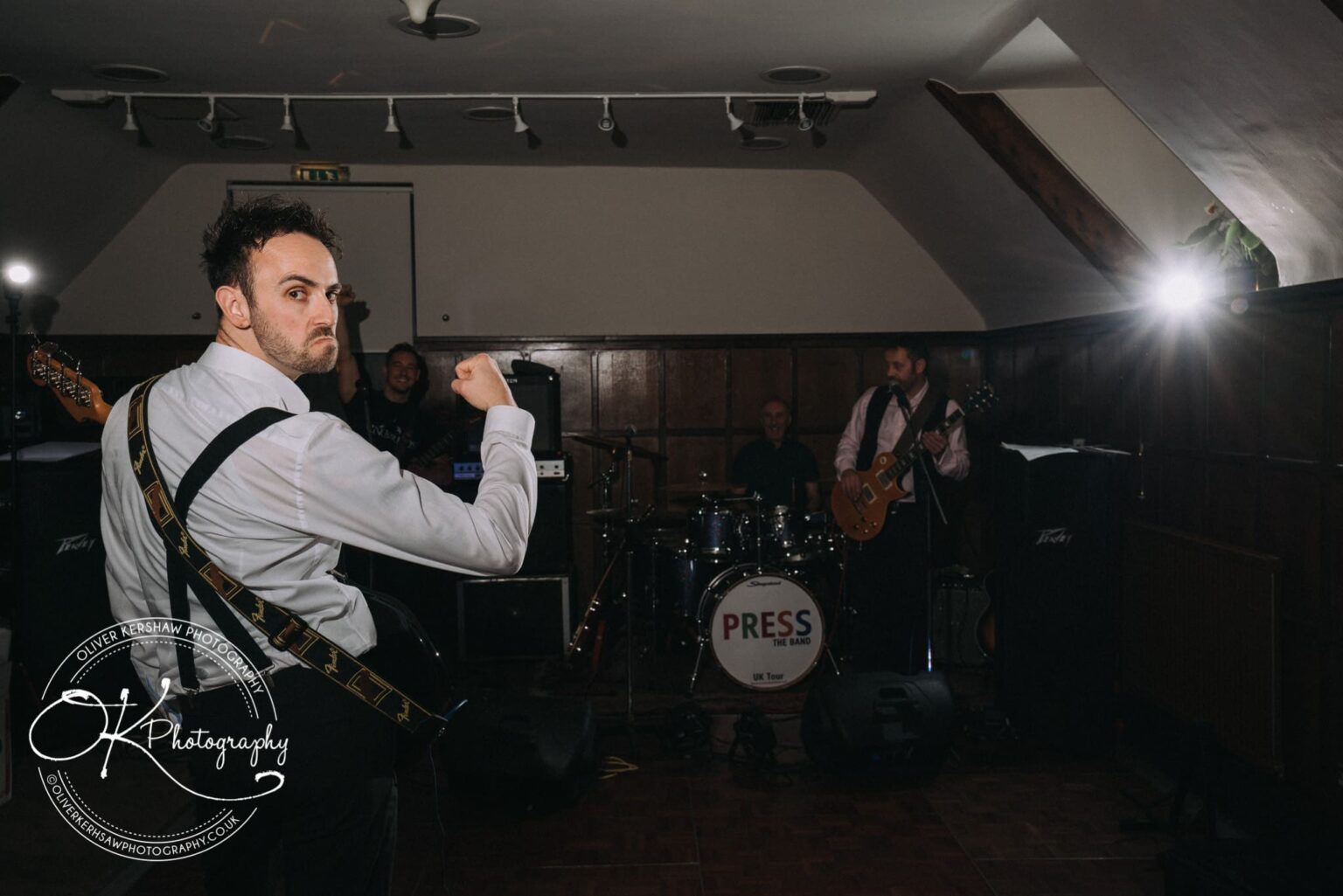 Wedding Photography George Hotel Stamford A band performing on a small stage, with a focus on a person in the foreground wearing a white shirt and holding a guitar, making a fist gesture. A drum set with "PRESS THE BAND" written on it is visible in the background.