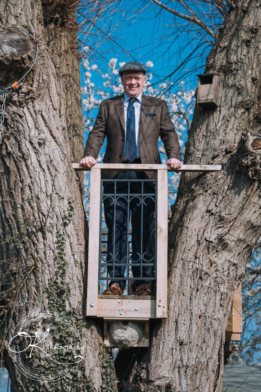 Wedding Photography George Hotel Stamford A man in a suit and flat cap stands on a wooden balcony built within a large tree, smiling at the camera.
