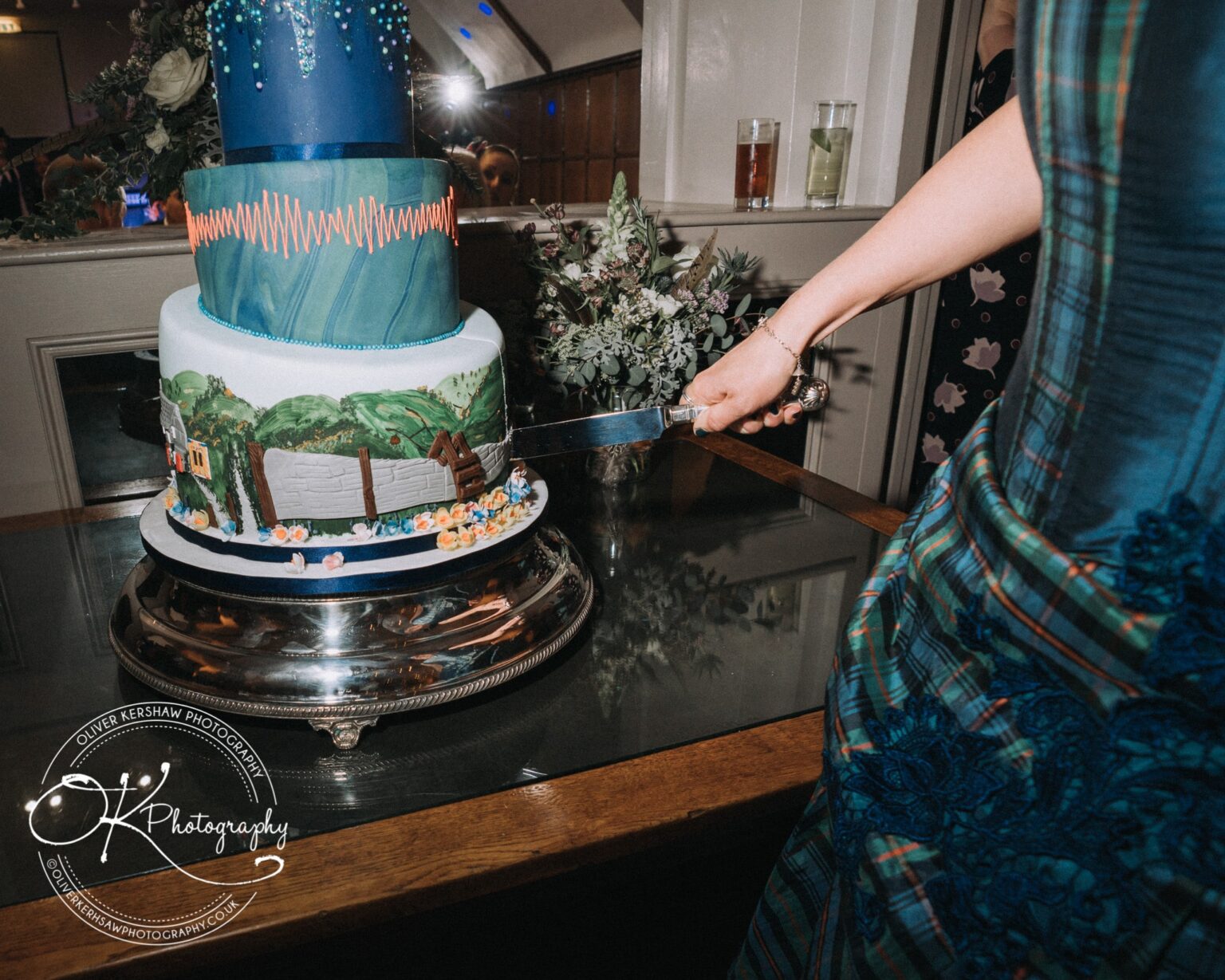 Wedding Photography George Hotel Stamford A person cutting a multi-tiered cake decorated with artistic designs, including a blue dripping effect on the top tier and a scenic illustration on the bottom tier. The cake is placed on a reflective silver stand on a wooden table.