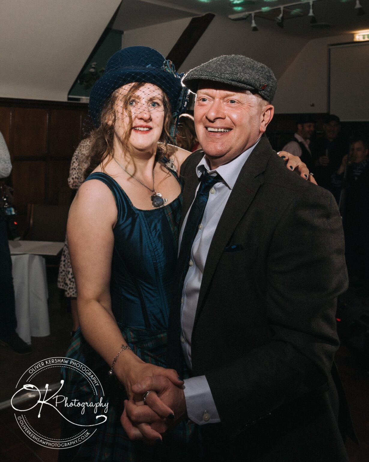 Wedding Photography George Hotel Stamford A smiling couple in formal attire dancing together, the woman wearing a blue dress and fascinator, and the man in a suit and flat cap.