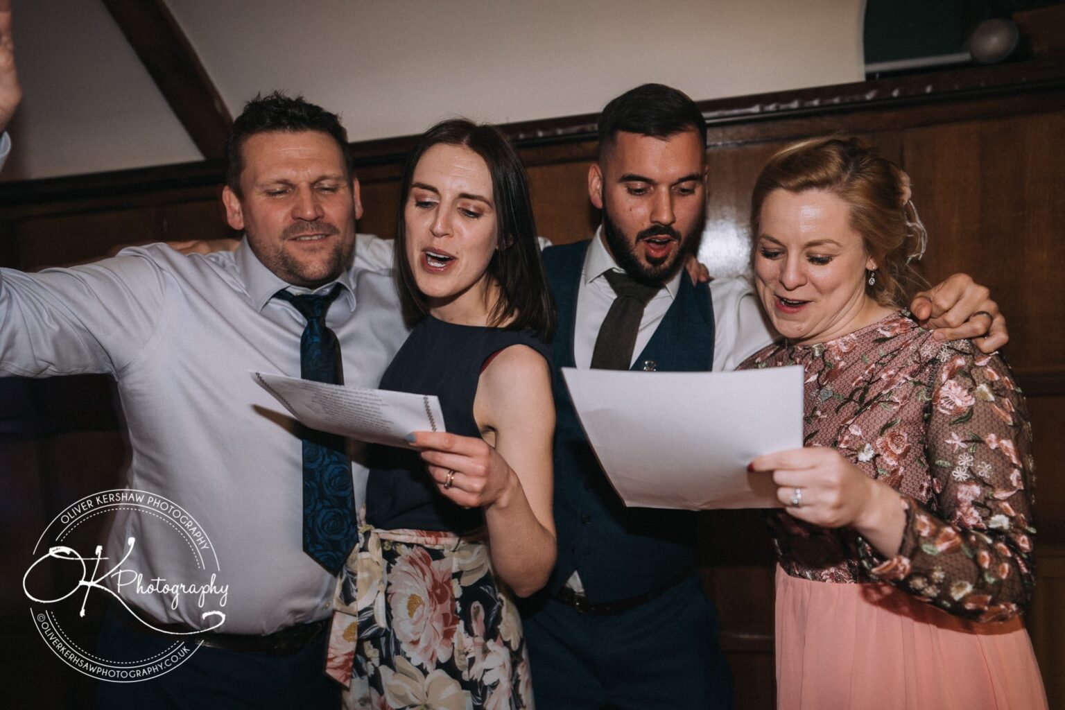 Wedding Photography George Hotel Stamford Four people sing together while holding papers, with one person raising their hand. The setting appears to be a formal or festive event.