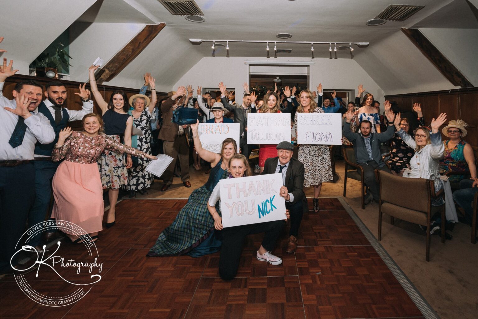 Wedding Photography George Hotel Stamford A group of people posing together in a room with wooden flooring, holding signs with names and "THANK YOU NICK," smiling and raising their hands.