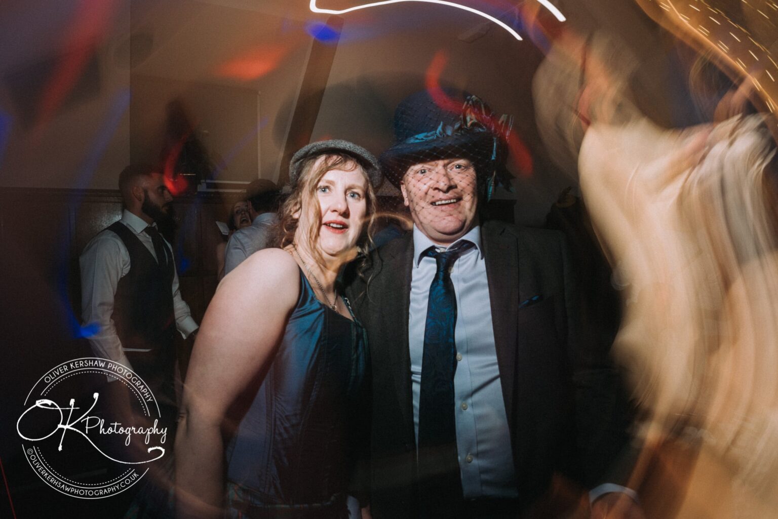 Wedding Photography George Hotel Stamford A couple wearing formal attire and vintage-style hats, posing for a photo at an indoor event with swirling lights and blurred background figures.