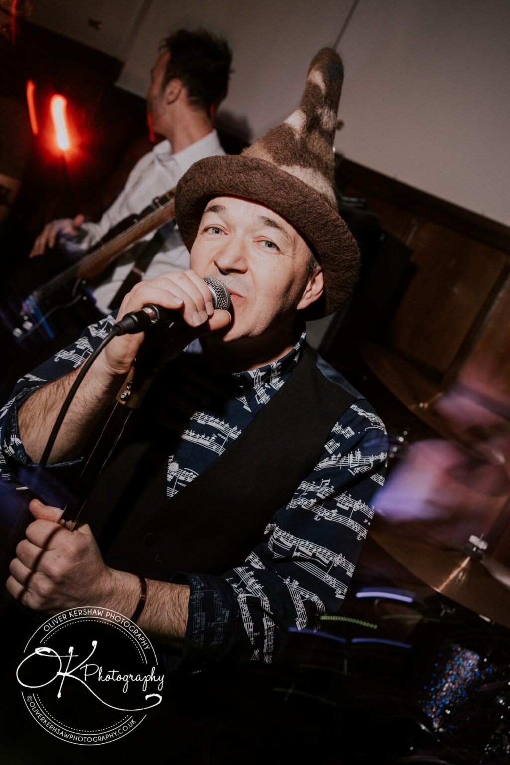 Wedding Photography George Hotel Stamford A musician wearing a unique hat and holding a microphone performs at George Hotel in Stamford. A second person playing an instrument is visible in the background.