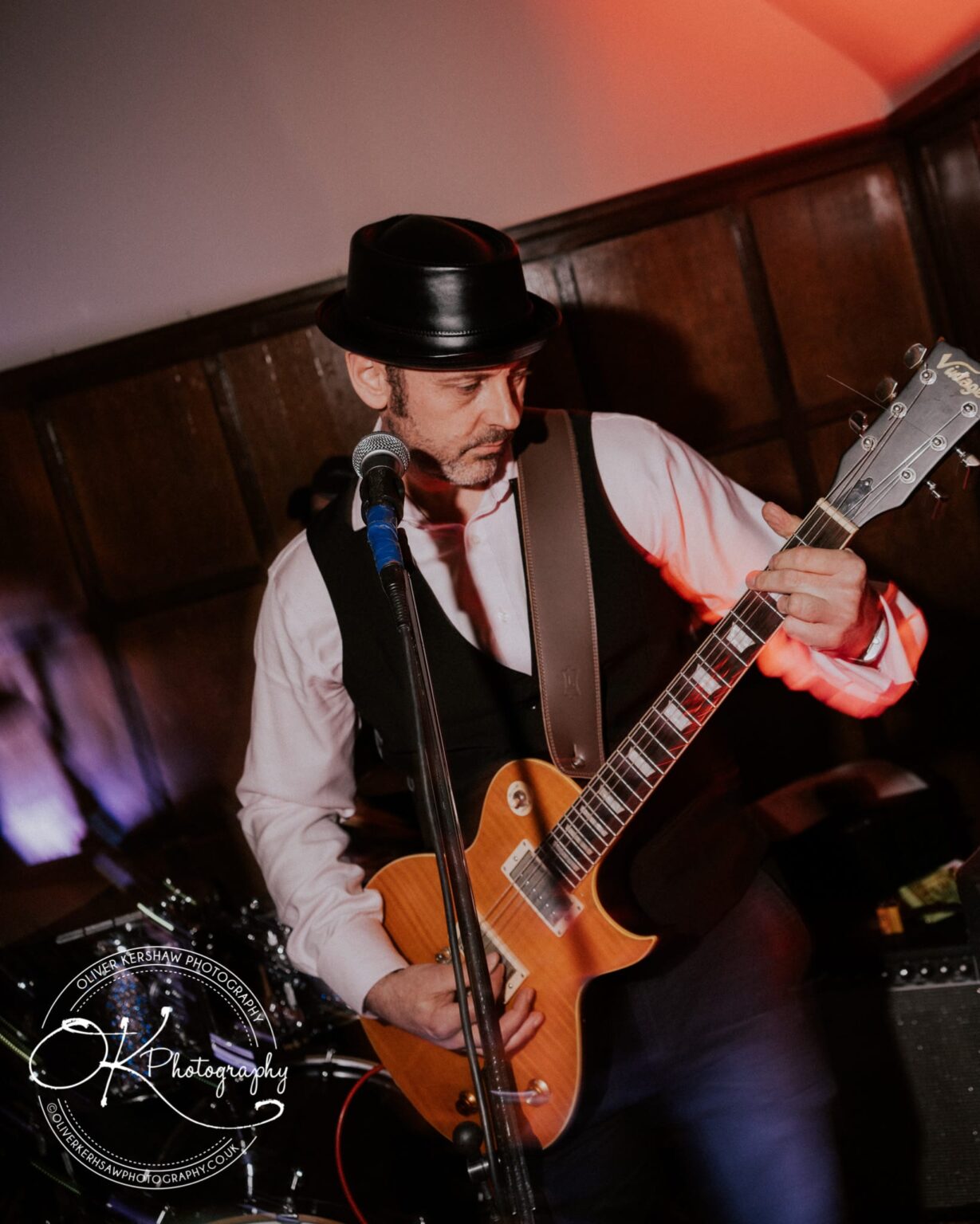 Wedding Photography George Hotel Stamford Man playing an electric guitar on stage in a formal indoor setting, wearing a hat, white shirt, and vest.