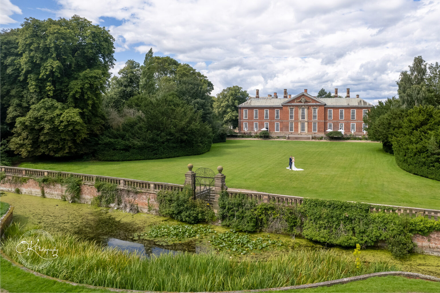 Aerial view of a couple in wedding attire standing on a vast green lawn in front of a large, historic brick mansion, surrounded by lush trees and greenery.