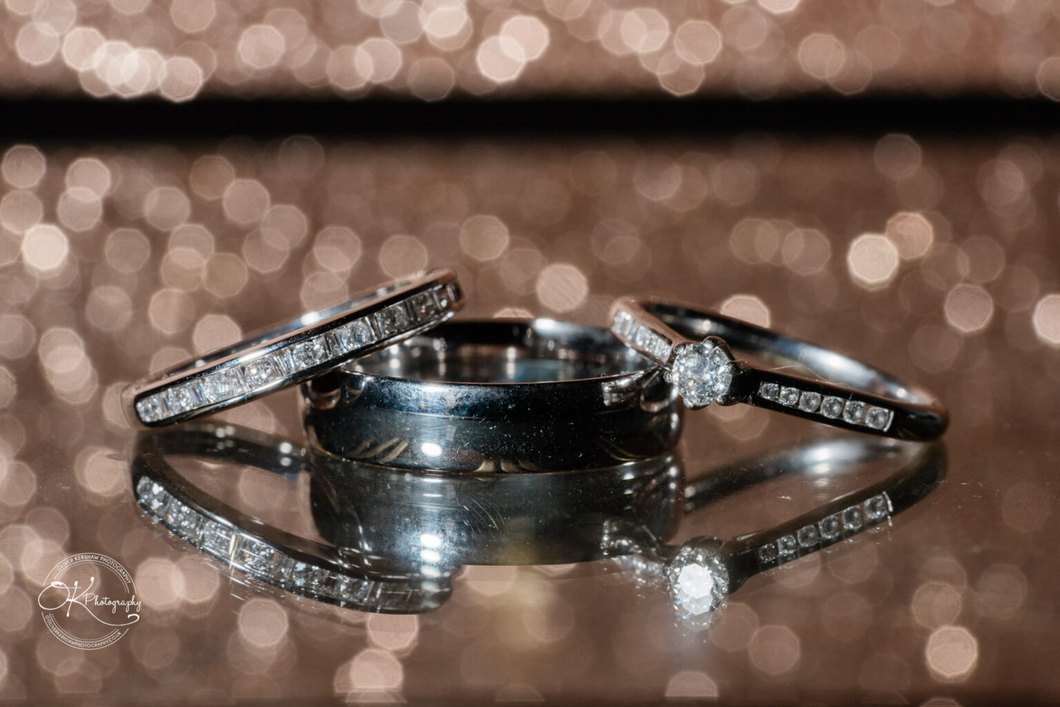 Three wedding rings, including one engagement ring with a diamond, photographed on a reflective surface with a bokeh background.