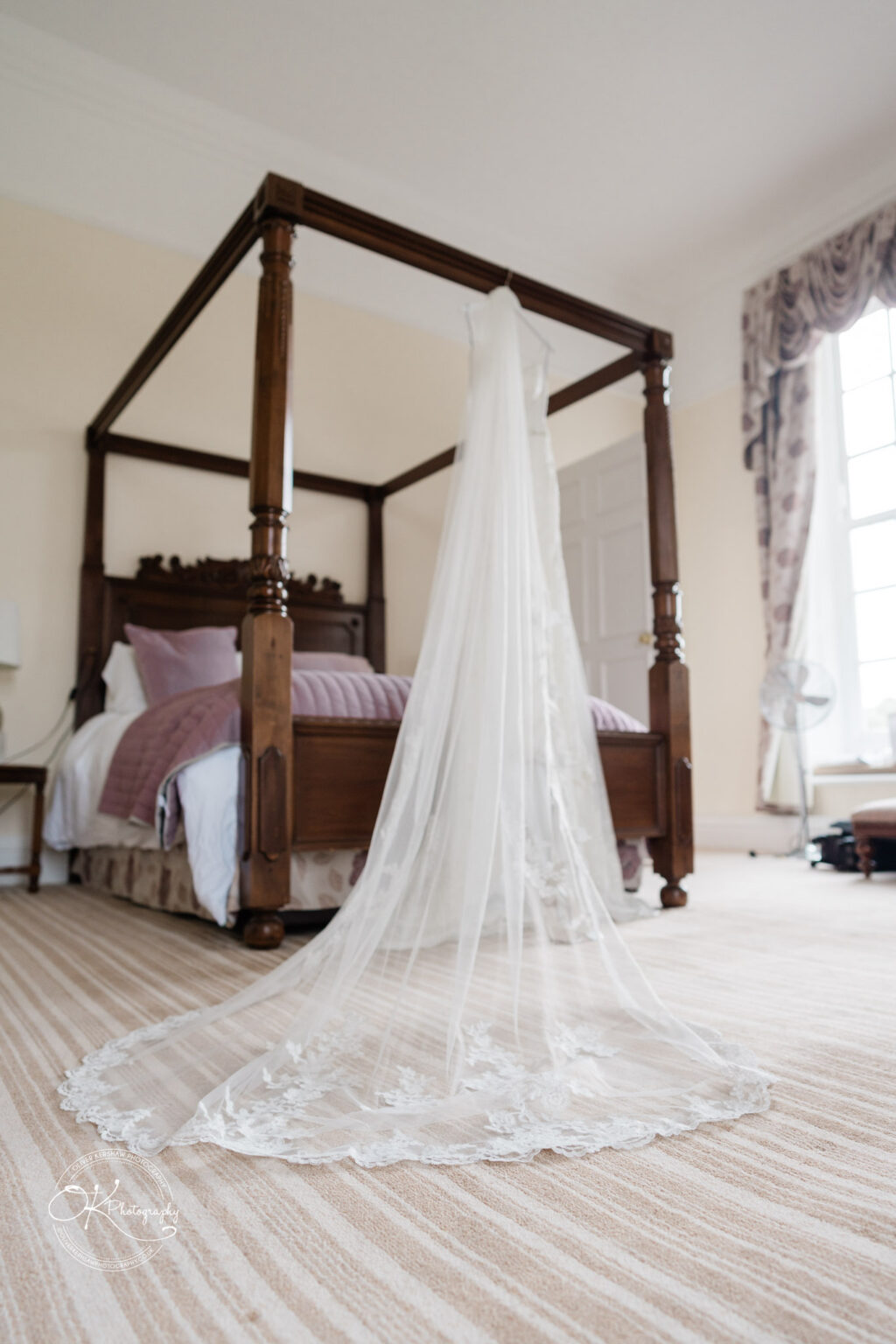A four-poster wooden bed in a hotel room, with a bridal veil draped across the top and trailing onto the floor. The room features a striped carpet, large window with floral curtains, and a bedside table with a lamp.