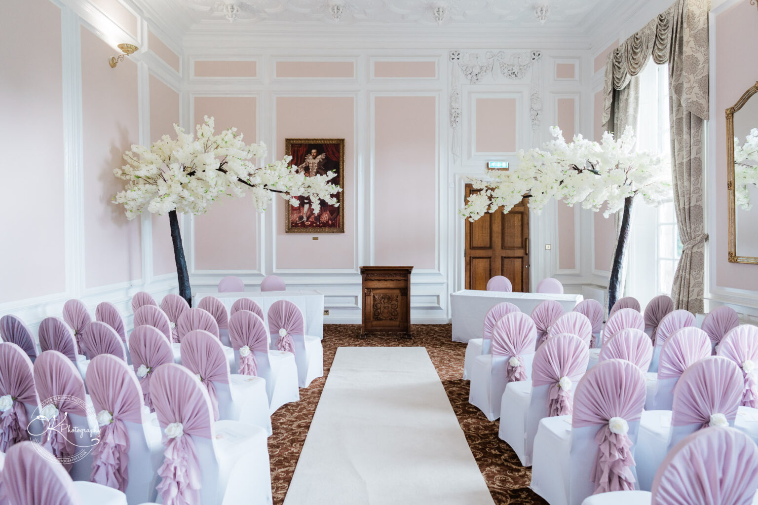 Elegant room set up for a wedding ceremony with rows of white chairs adorned with lavender fabric, a white aisle runner, and artificial white cherry blossom trees.