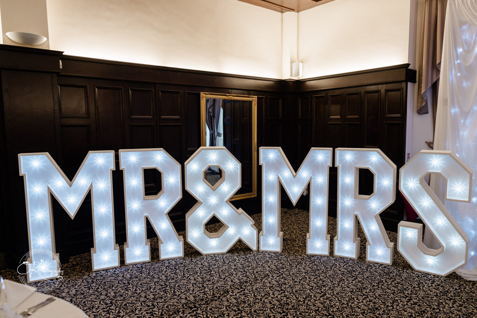 Large illuminated letters spelling "MR&MRS" in a room with dark wood panelling and a patterned carpet at Bosworth Hall Hotel.