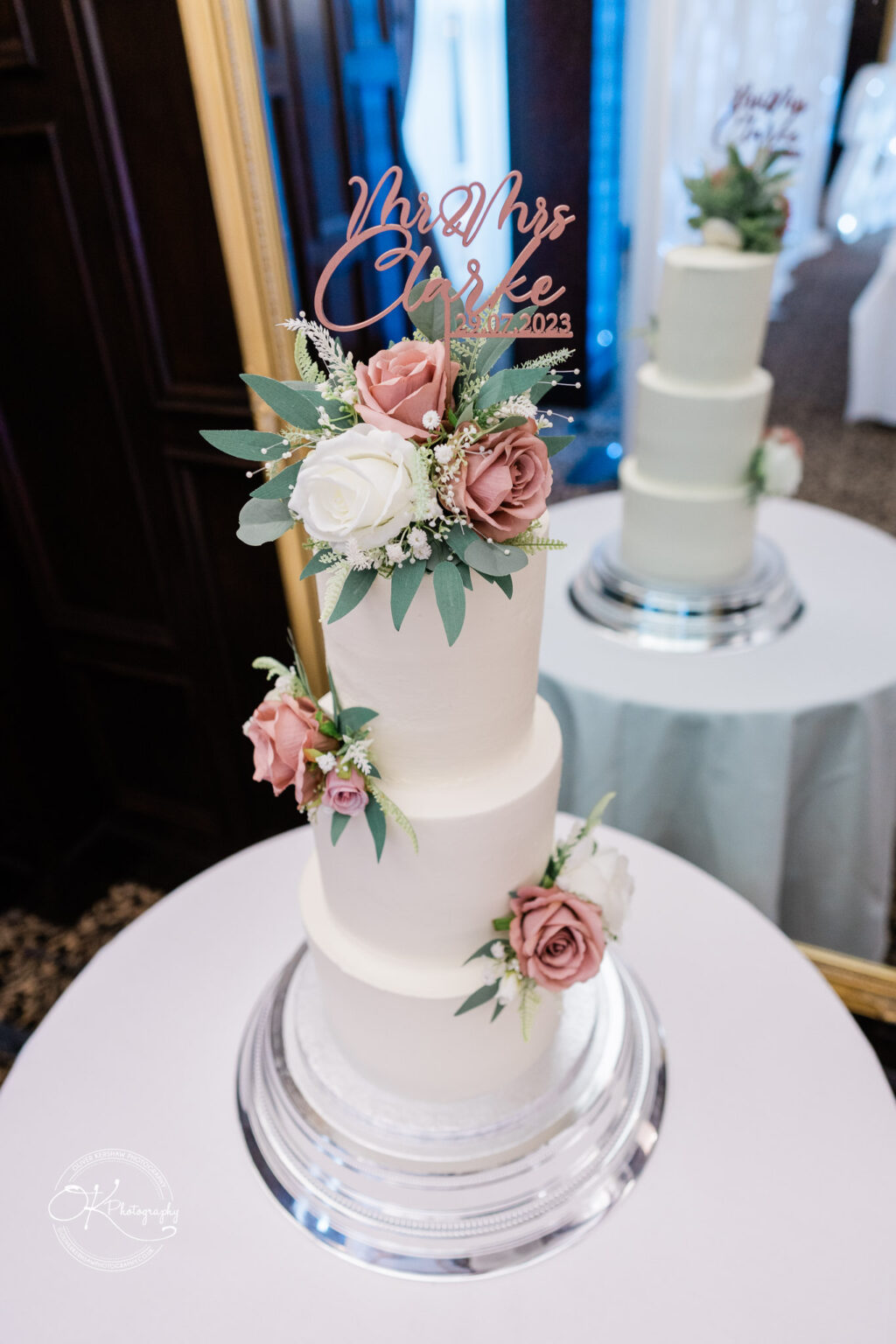 A white, three-tiered wedding cake decorated with roses and greenery, topped with a plaque reading "Mr & Mrs Clarke 21.07.2023," displayed on a silver cake stand.