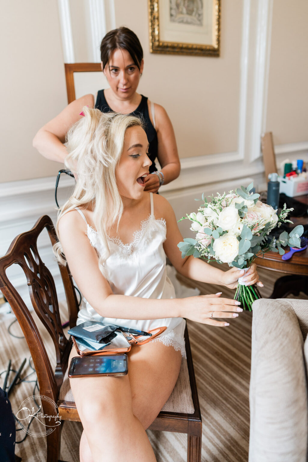 A woman in a silk camisole holding a bouquet, having her hair styled by another woman in a room with ornate details and wooden furniture.