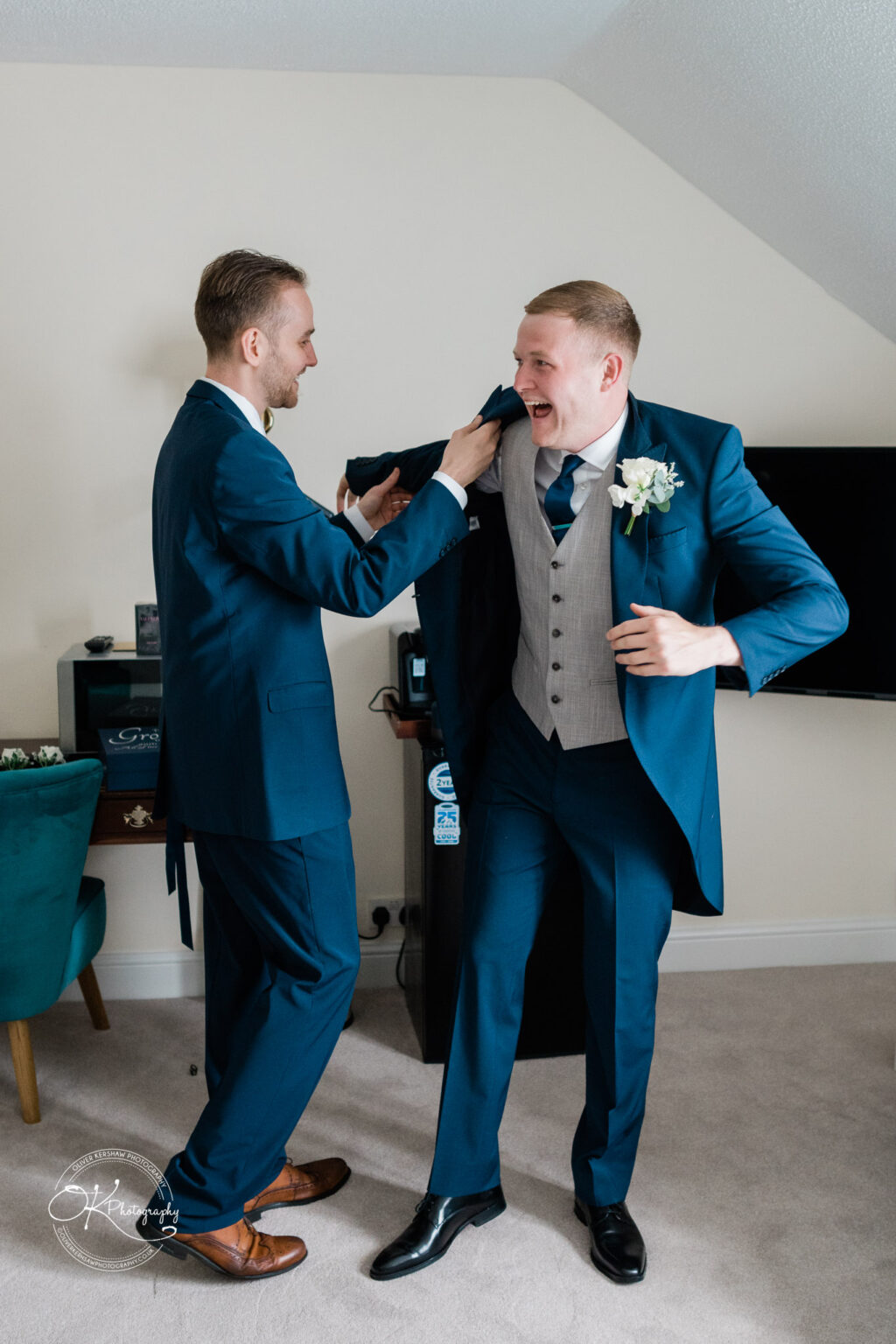 Two men in blue suits and waistcoats enjoy a light-hearted moment while getting ready in a hotel room, one helping the other with his jacket.