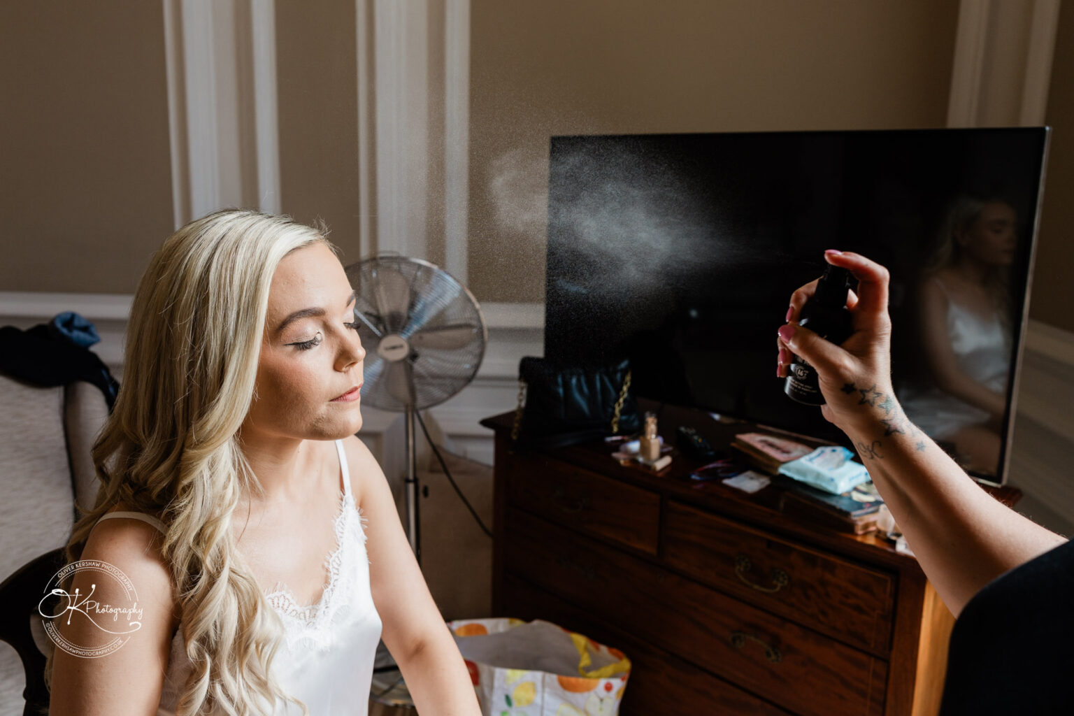 A woman having her hair styled with hairspray in a room, with a fan, a TV, and a wooden dresser in the background.