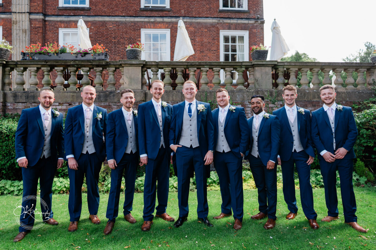 A group of men in matching blue suits and waistcoats posing in front of a brick building with a stone balustrade and flower arrangements.