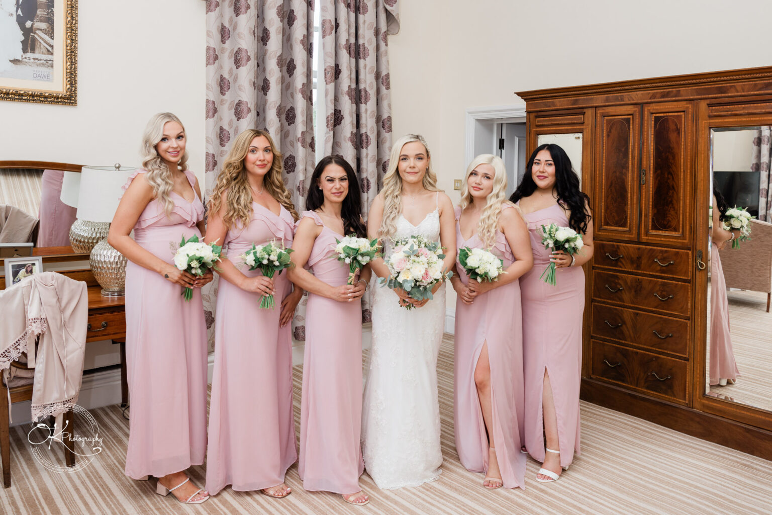 A bride and her bridesmaids dressed in light pink gowns stand in a room at Bosworth Hall Hotel, holding bouquets of white flowers.