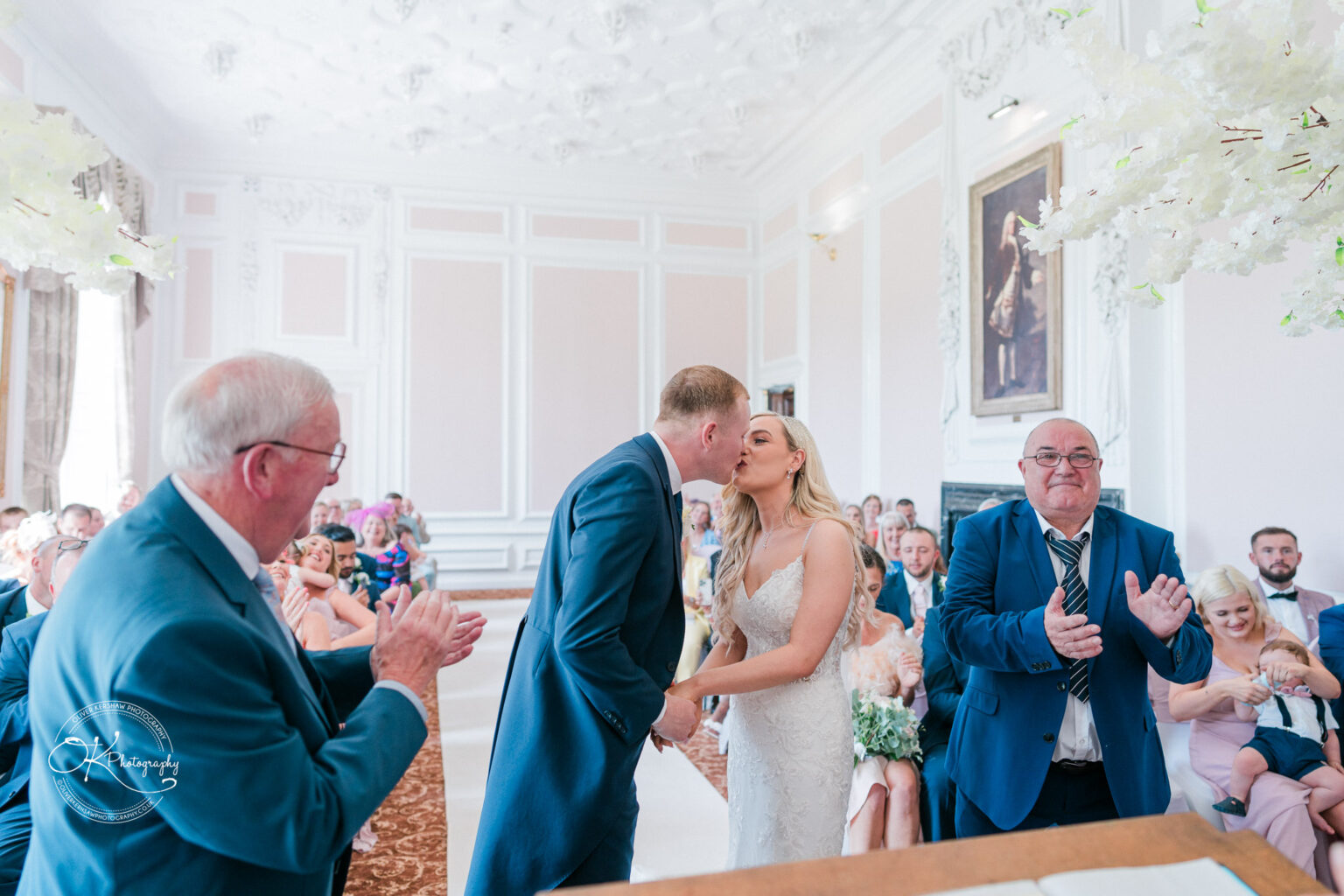 Bride and groom kissing during a wedding ceremony at Bosworth Hall Hotel, with the guests seated and applauding.