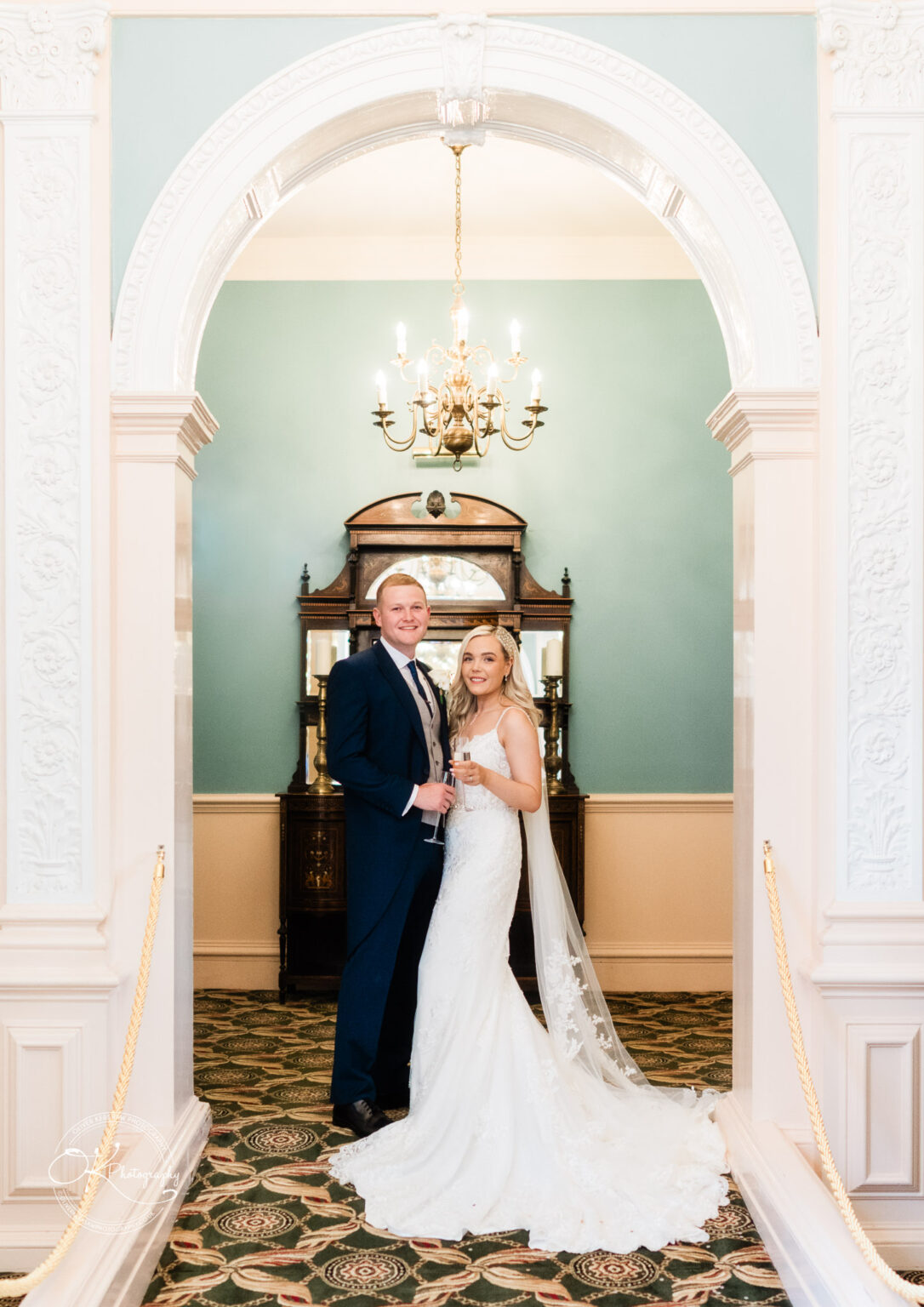A bride and groom standing under an ornate archway at Bosworth Hall Hotel, lit by a chandelier, with the bride in a white wedding dress and the groom in a blue suit.