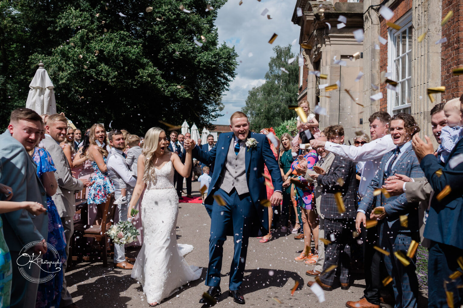 Bride and groom walking down the aisle at an outdoor wedding ceremony, surrounded by cheering guests throwing confetti.
