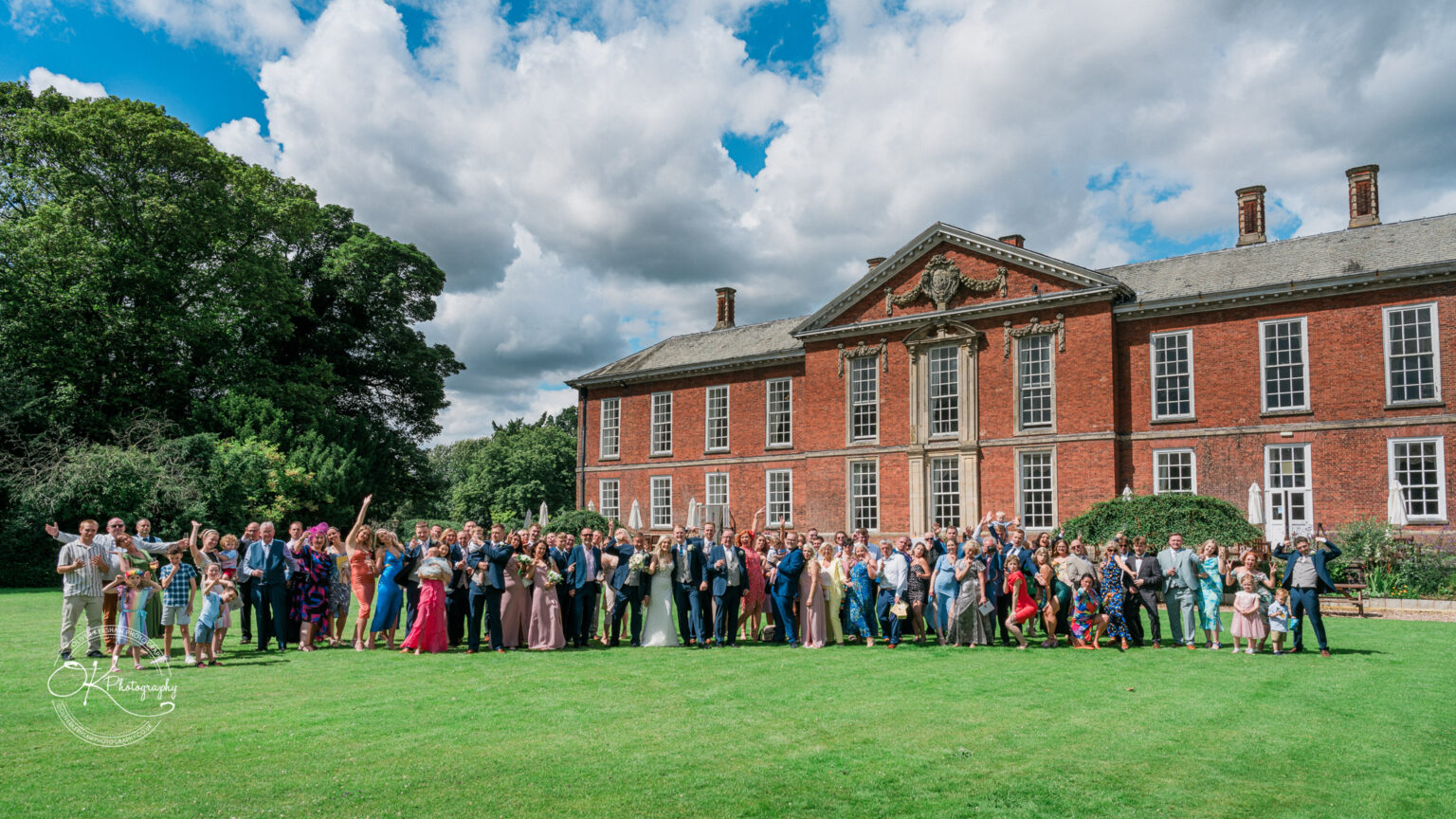 A large group of people posing on a landscaped lawn in front of Bosworth Hall Hotel, a grand red-brick building with ornate architectural details and multiple windows.