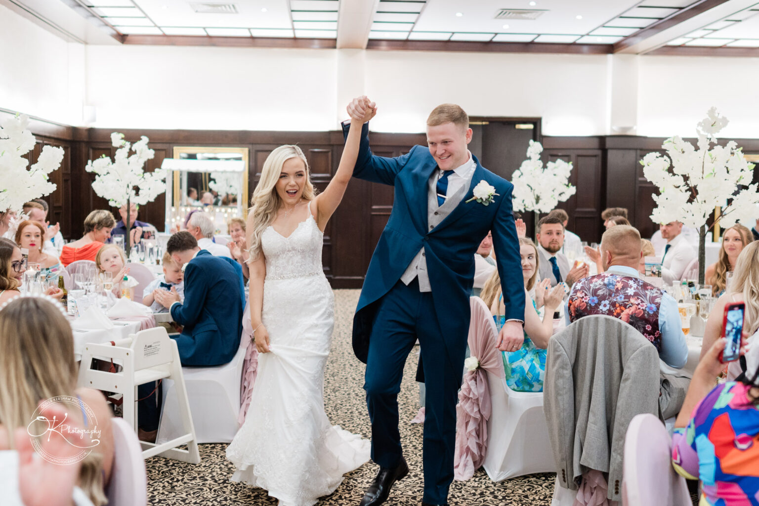 A bride and groom celebrating in a banquet hall with seated guests clapping and cheering.