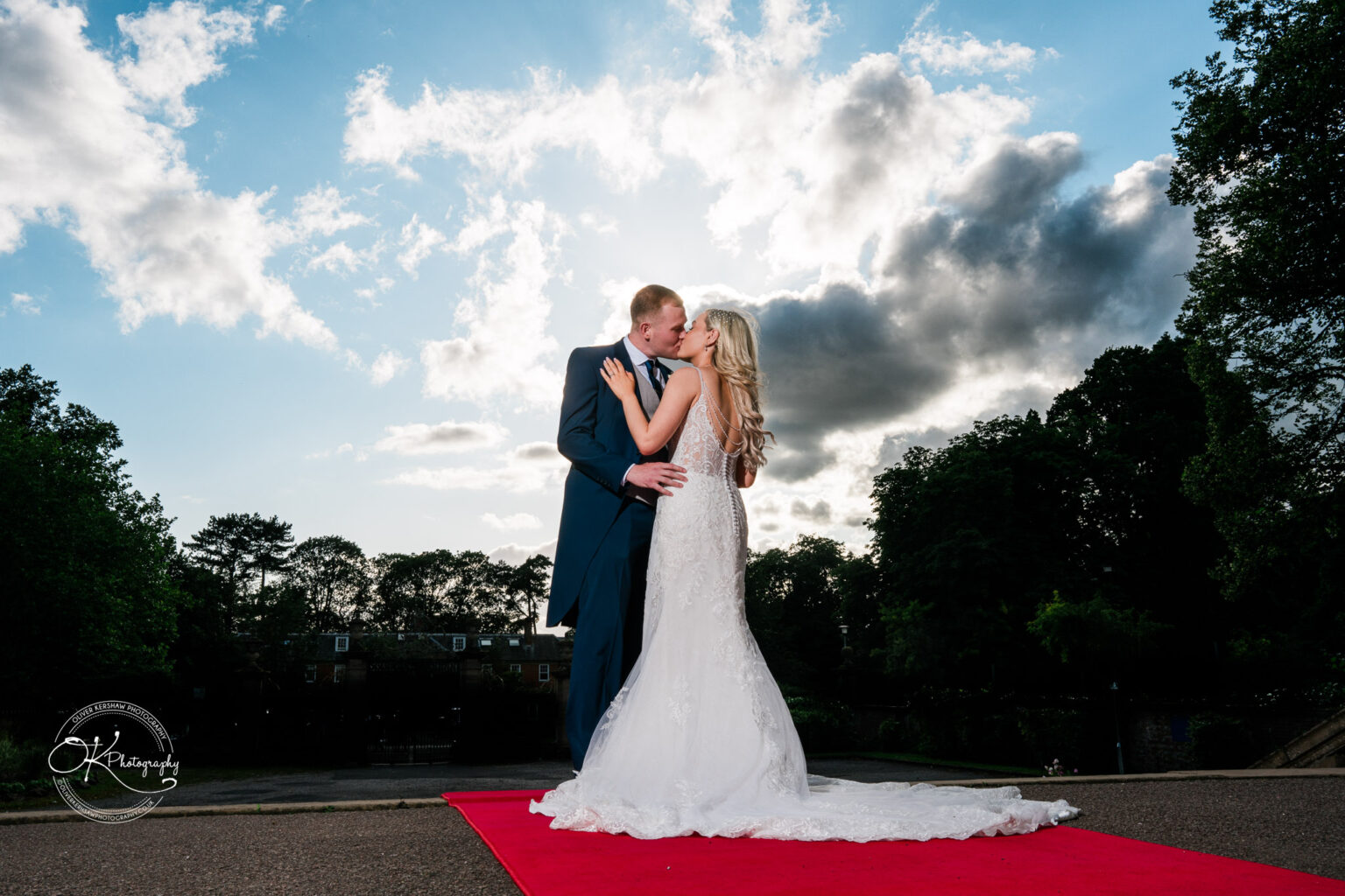 A newlywed couple kissing on a red carpet outdoors, with a dramatic cloudy sky and trees in the background.