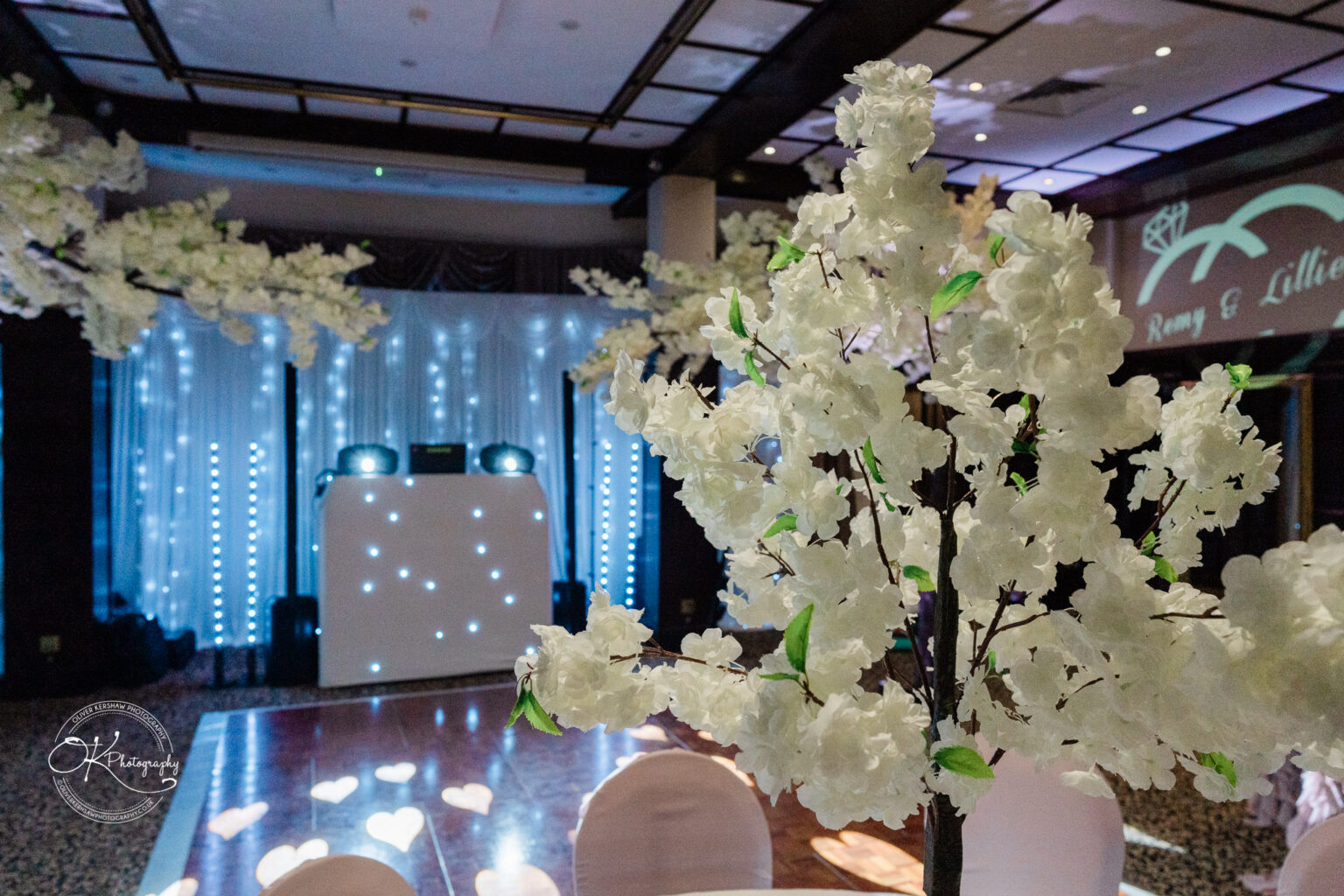 Wedding reception hall decorated with white flowers, a lit dance floor, and a DJ booth with lights.