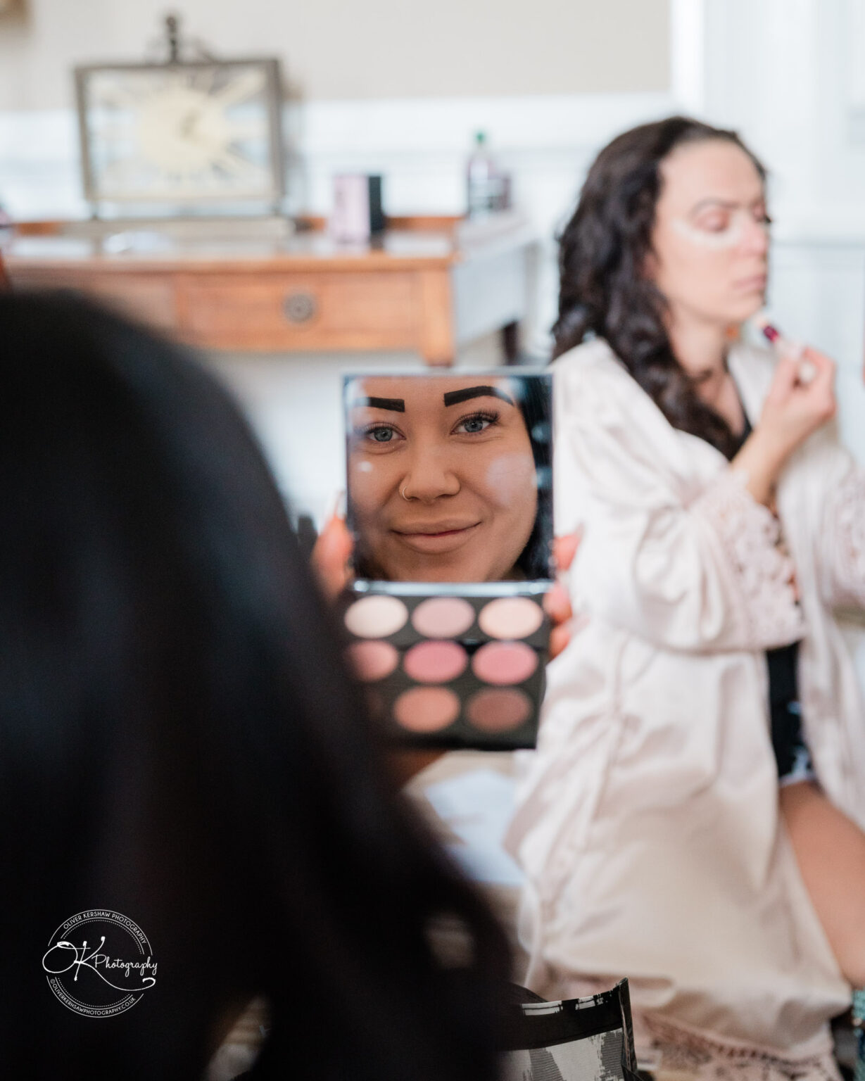 A woman holding a makeup mirror reflecting her face, with another woman in the background applying lipstick.