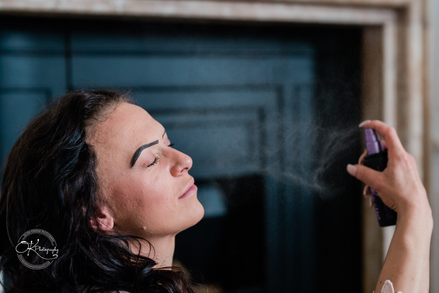 A person with closed eyes spraying mist on their face from a bottle, with "OK Photography" watermark in the bottom left corner.