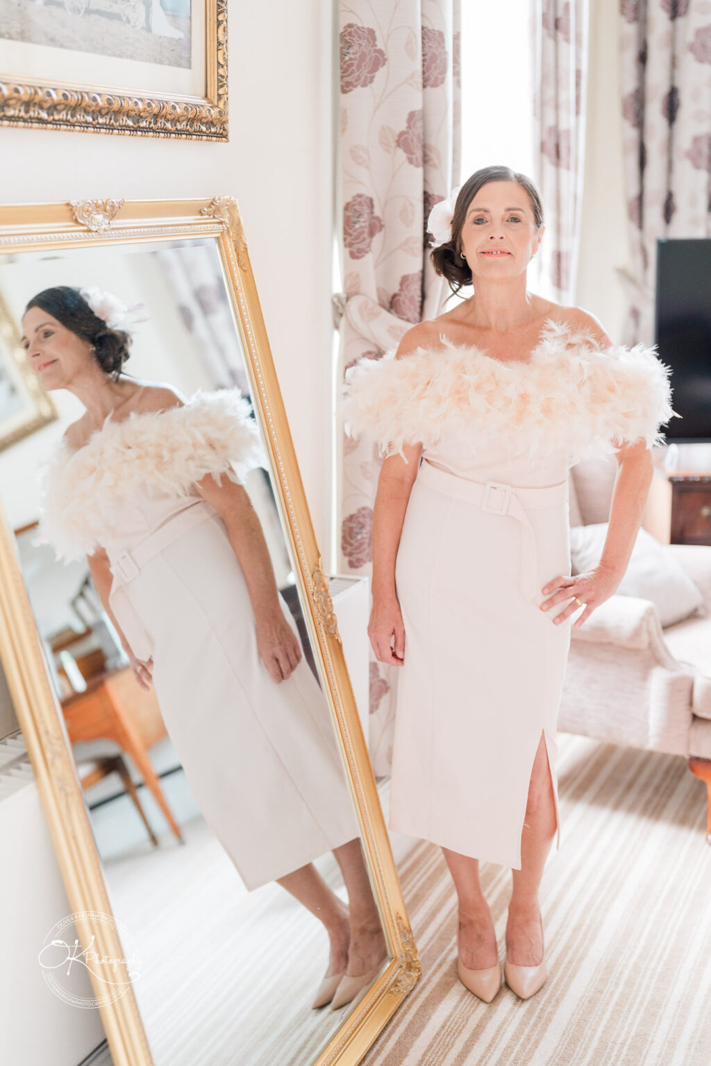 Woman in an elegant off-shoulder dress with feather detail, standing next to a large gold framed mirror in a tastefully decorated room with floral curtains.