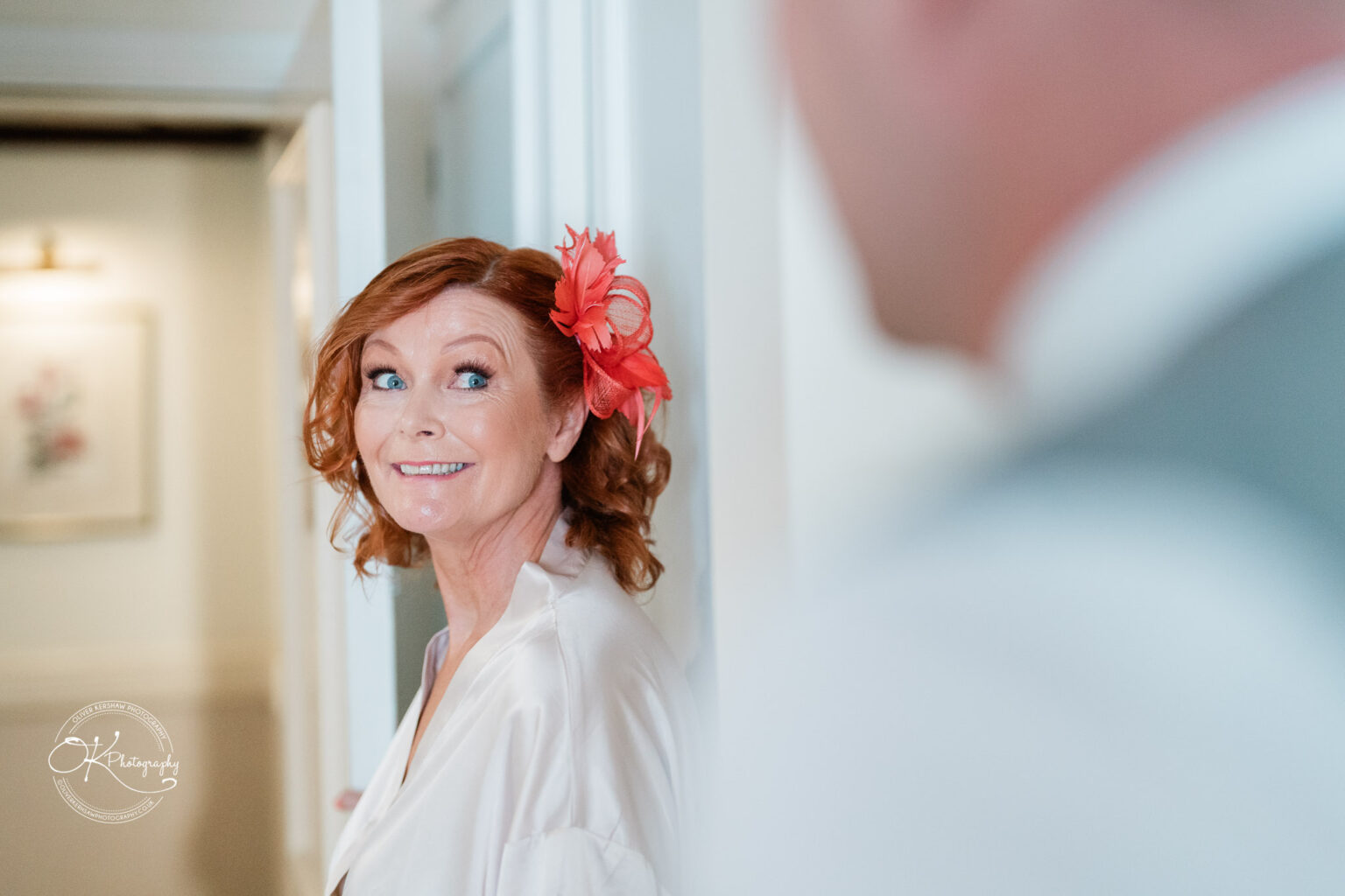 A woman with red hair and a red fascinator smiles while looking at someone off-camera.