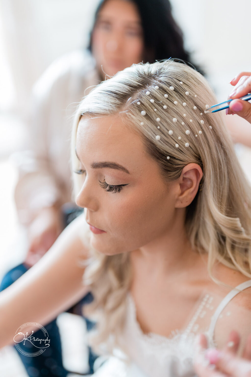 A close-up of a woman having small white pearls placed in her blonde hair, with another woman in the blurred background.