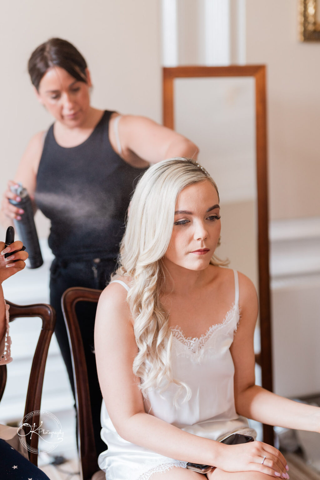 A woman with long, curled blonde hair sitting in a chair, while another woman in a black tank top uses hairspray on her hair.