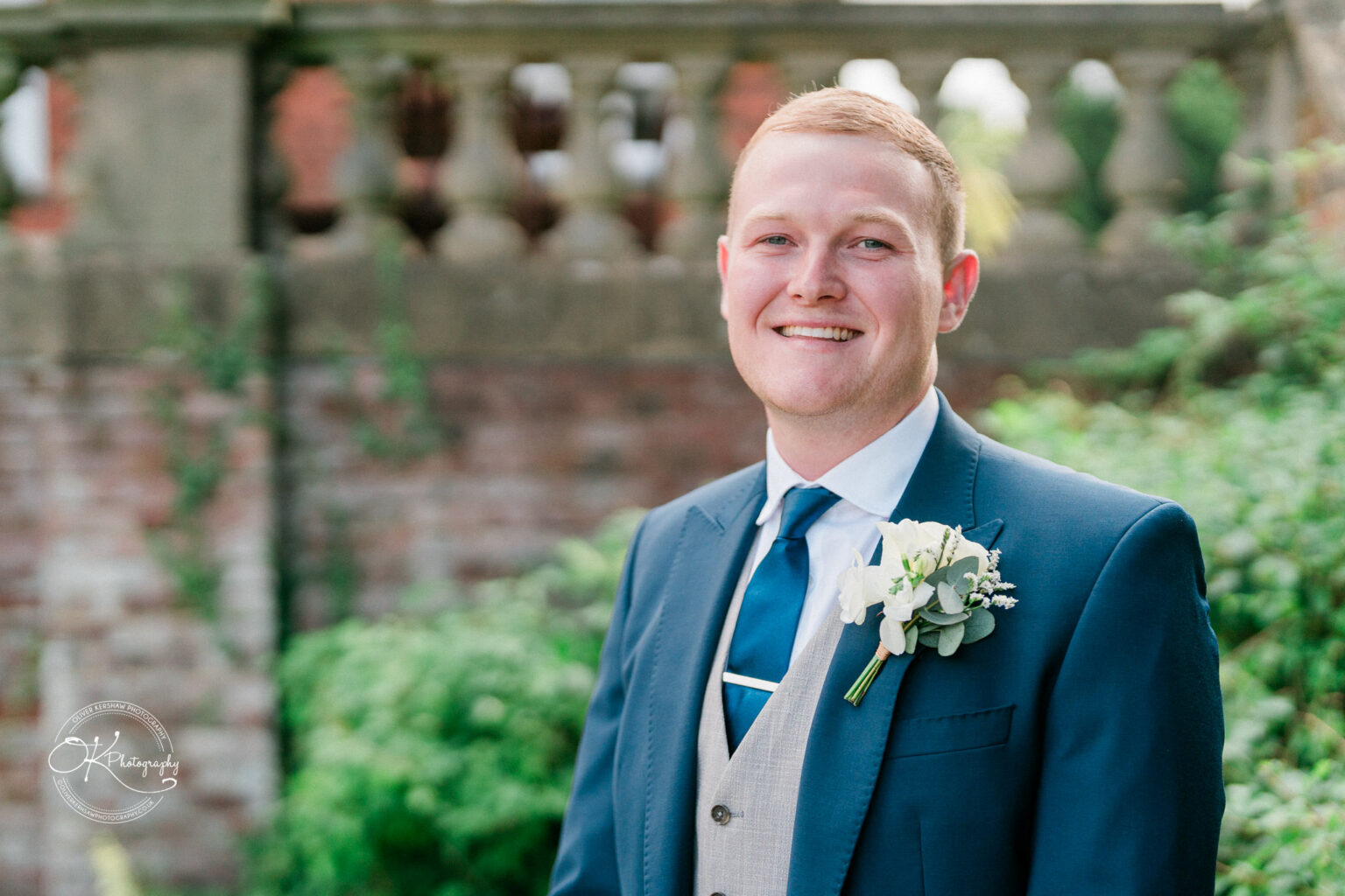 Man in a blue suit and gray waistcoat with boutonnière, standing outdoors and smiling.