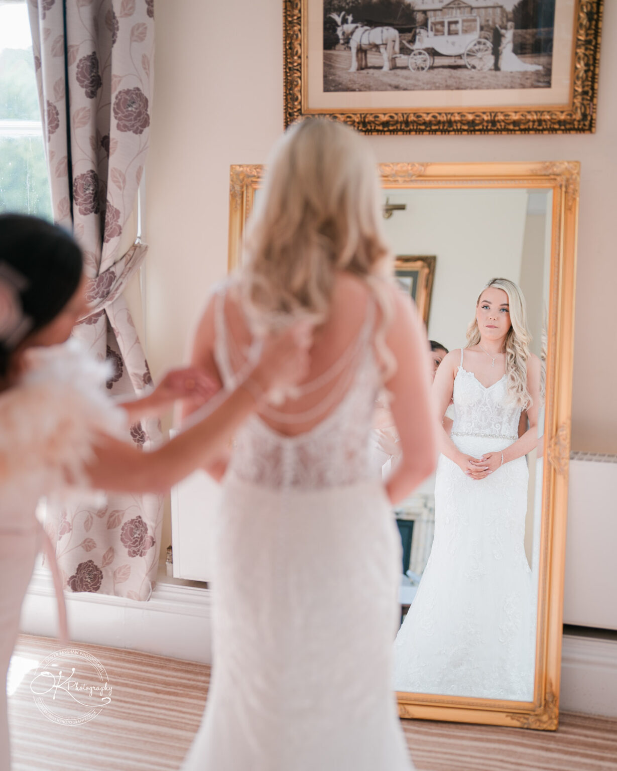Bride in wedding dress stands in front of a mirror being assisted by another person.