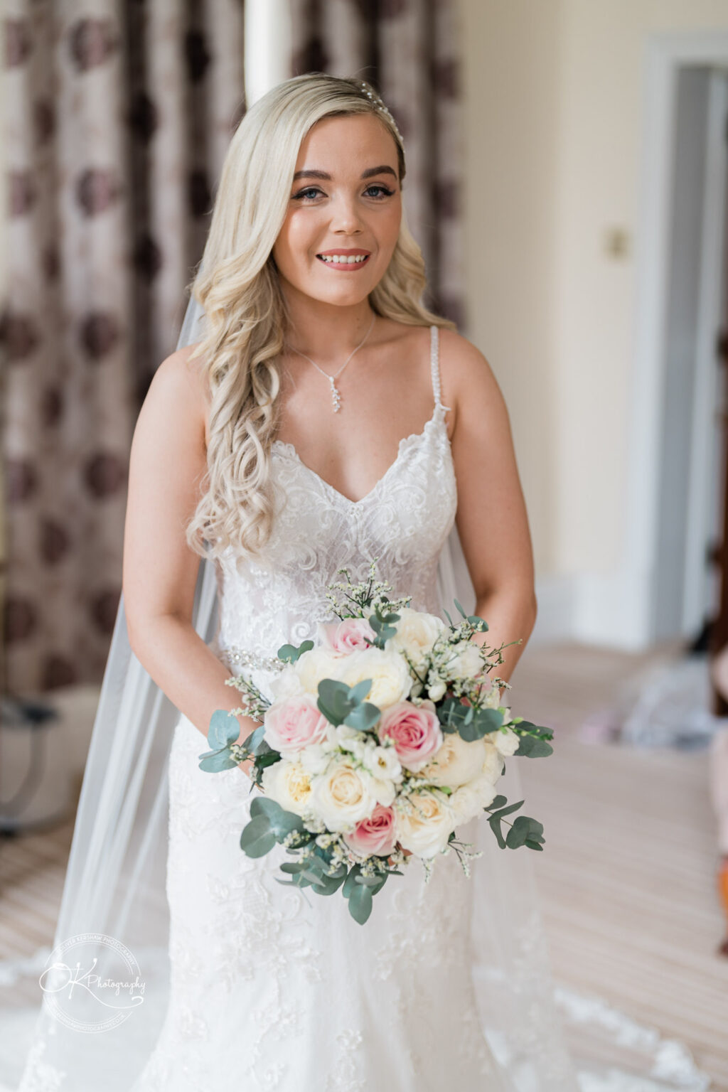 A bride in a lace wedding dress holds a bouquet of white and pink flowers, standing indoors with light-coloured walls and patterned curtains in the background.