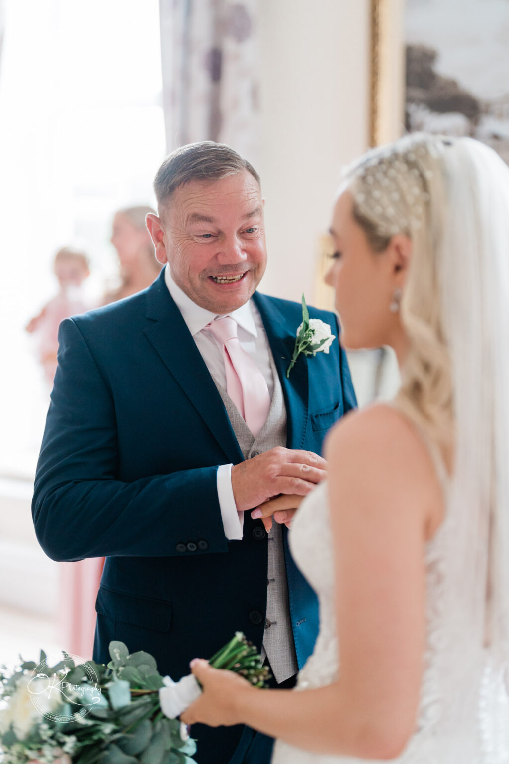 A man in a blue suit smiling and holding hands with a woman in a wedding dress, indoors.