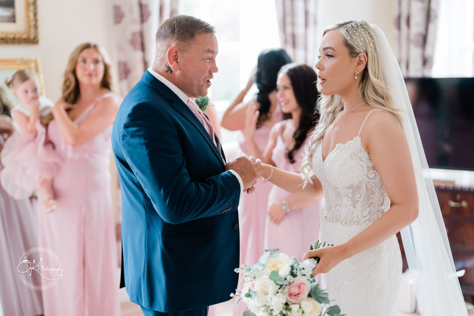 A bride in a lace wedding dress holding a bouquet is speaking with a man in a blue suit. Bridesmaids in pink dresses are in the background.