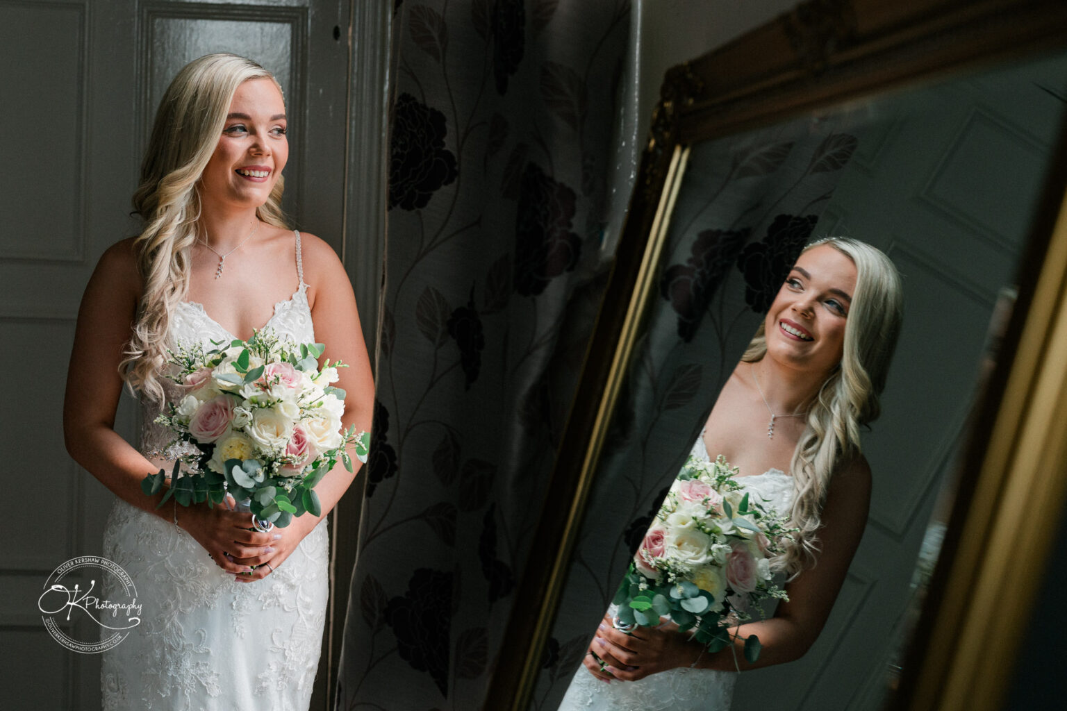 Bride in a white lace gown holding a bouquet, smiling at her reflection in a mirror.
